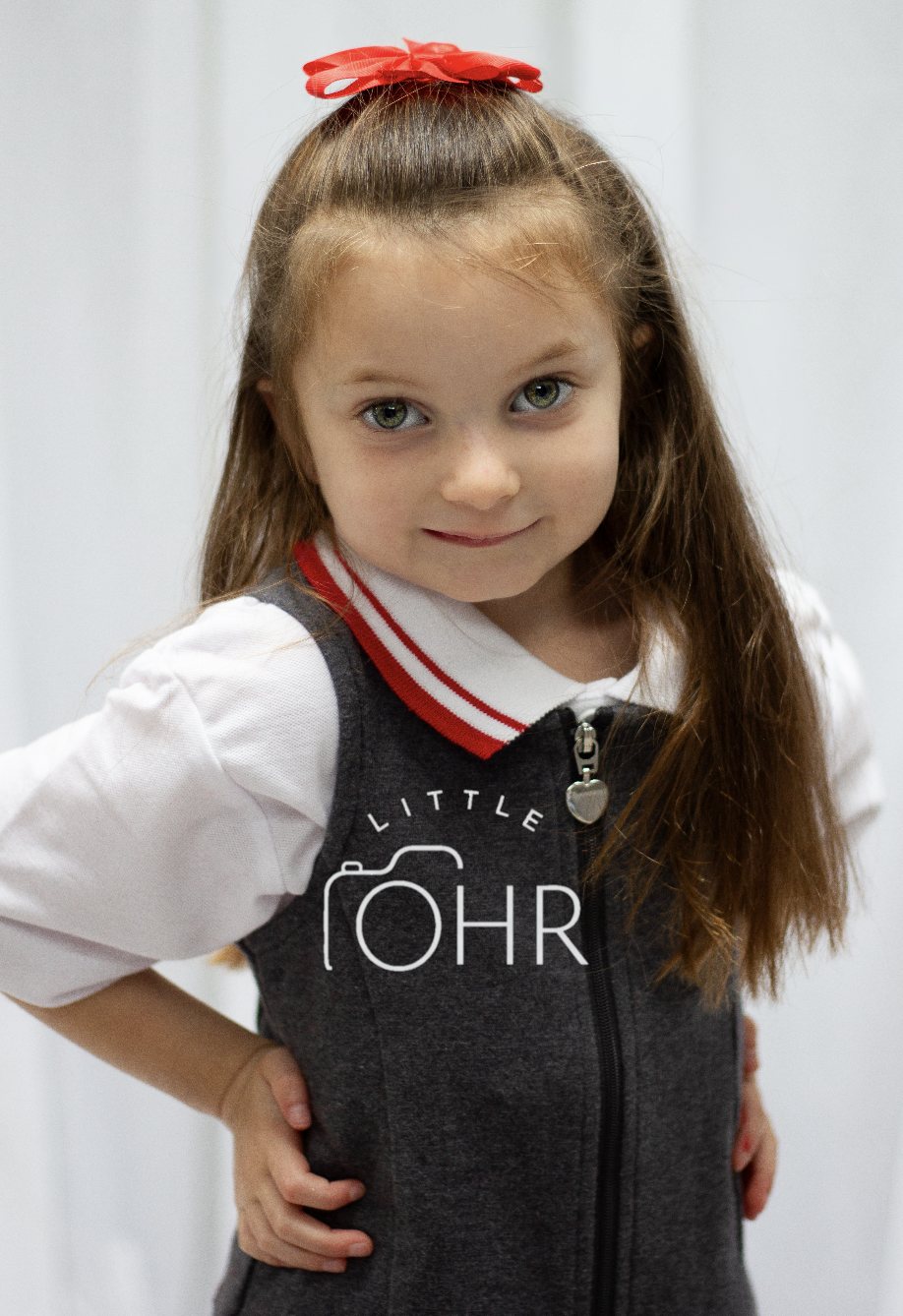 Girl with a red bow in her hair, hands on hips, in a grey school pinafore